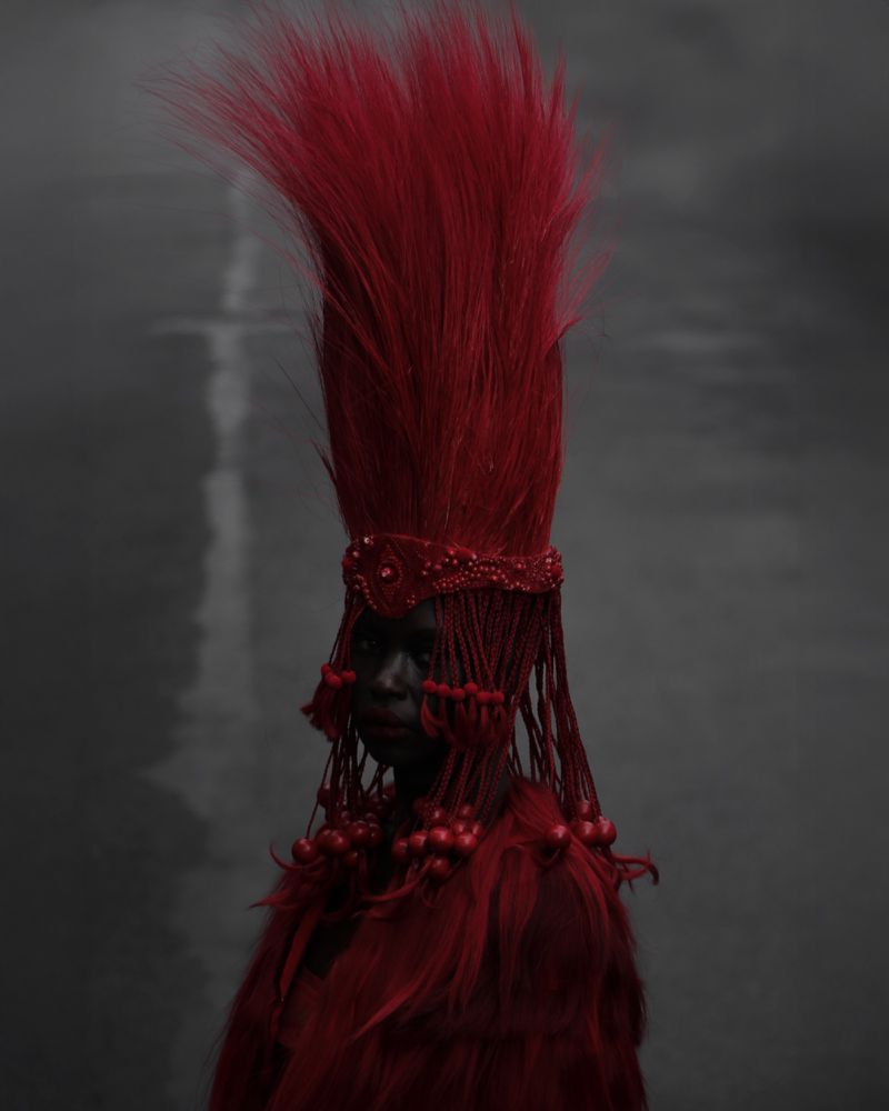 Close-up portrait of person with dark skin, elaborate red headdress and beaded collar against a gray background with light reflection.