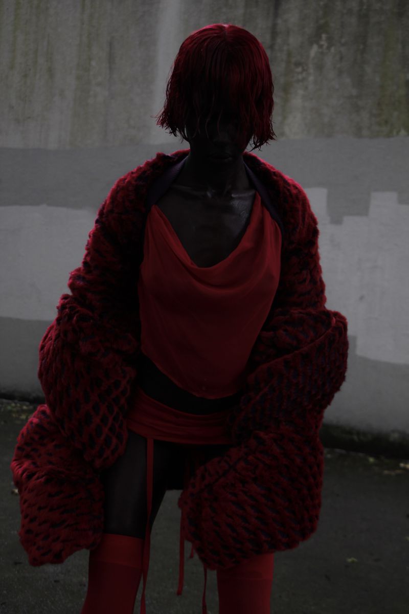 A full-length shot presents a woman wearing a red wig, a red top, and a large knitted red sweater, posing against a grey wall outdoors.