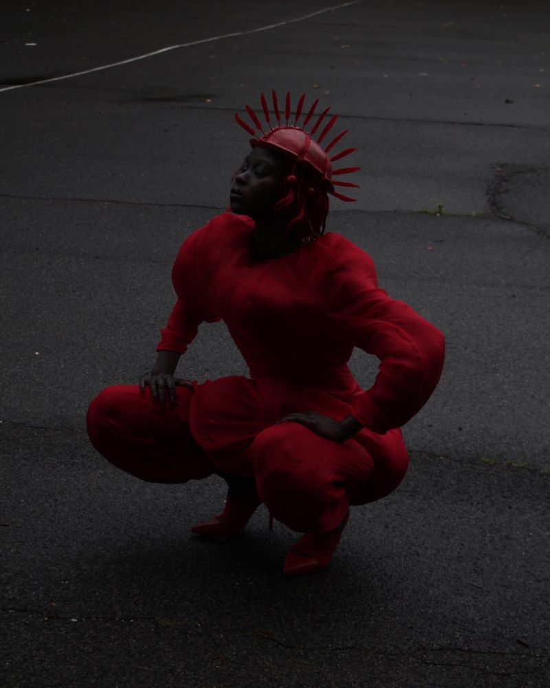 Person in red costume with headpiece squats on a paved surface in dimly lit outdoor shot, creating a striking monochromatic visual.