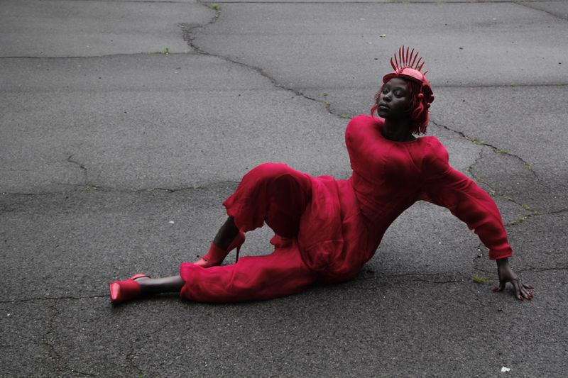 A woman in a red outfit and headpiece reclines on cracked asphalt, creating a striking fashion portrait with a monochromatic color scheme.