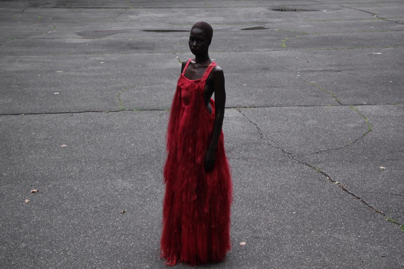A solitary figure stands outdoors, a woman in a textured red dress, captured in a medium shot with a slightly overcast aesthetic.