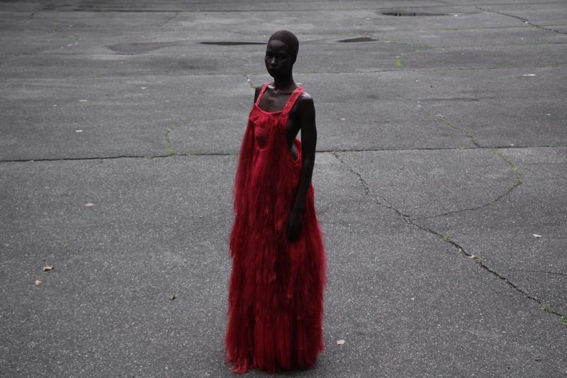 A solitary figure stands outdoors, a woman in a textured red dress, captured in a medium shot with a slightly overcast aesthetic.