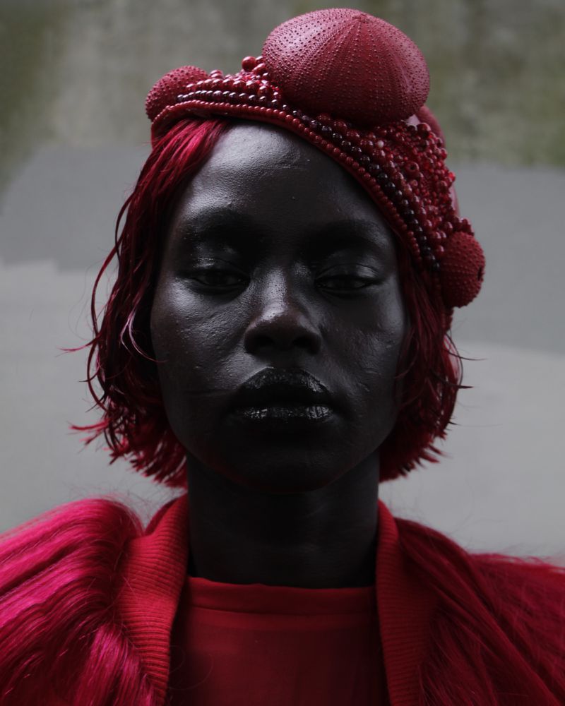 Close-up portrait of a black woman with red hair and artistic red beaded headwear, glossy black lips, and a red outfit in a studio.