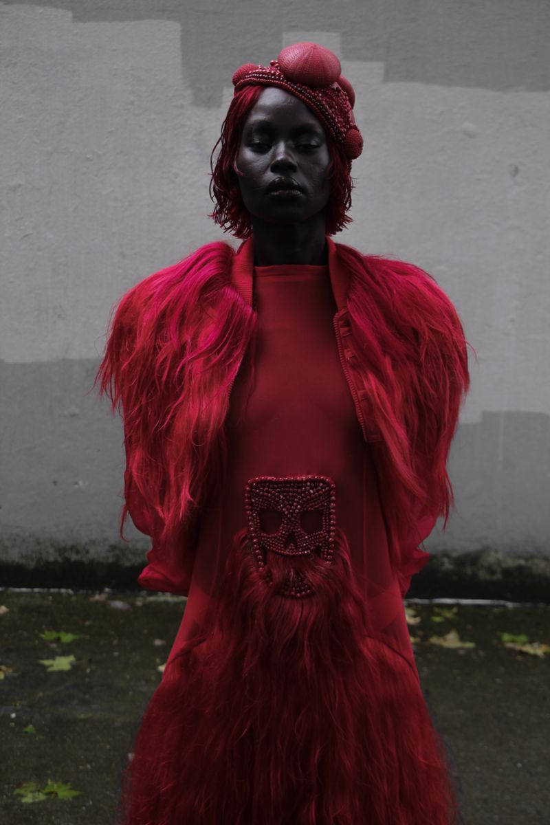 Fashionable woman with striking red hair, dressed in a vibrant red dress with fur-like detailing, poses against a textured gray wall.