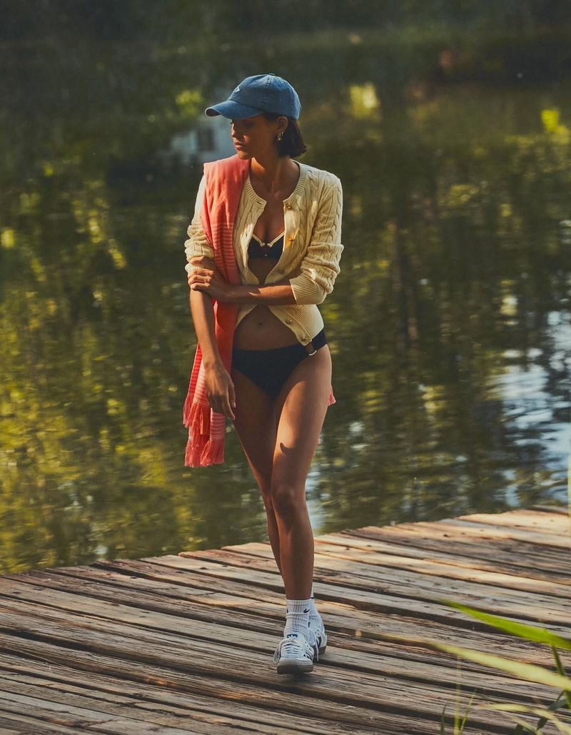 A young woman in a bikini stands on a wooden dock near a lake, wearing a sweater and baseball cap, enjoying a sunny, relaxed day.
