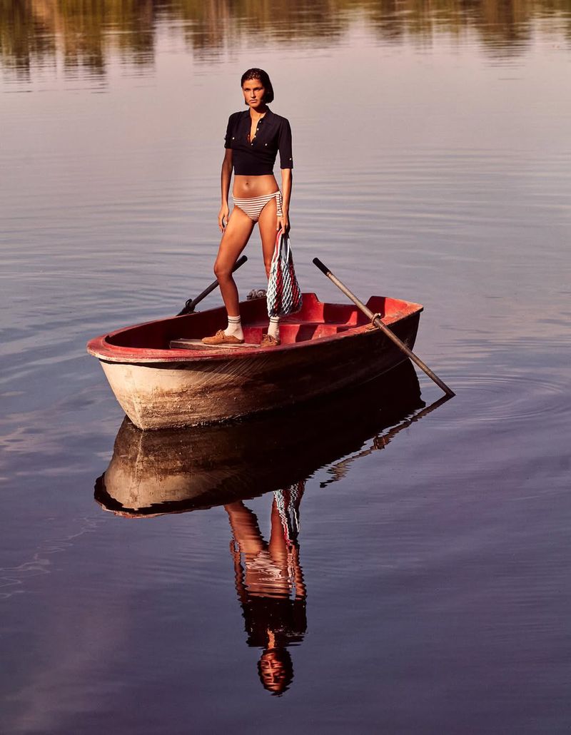 Woman in bikini and shirt standing on boat in calm water with reflection, holding bag and oars with natural setting, medium shot.
