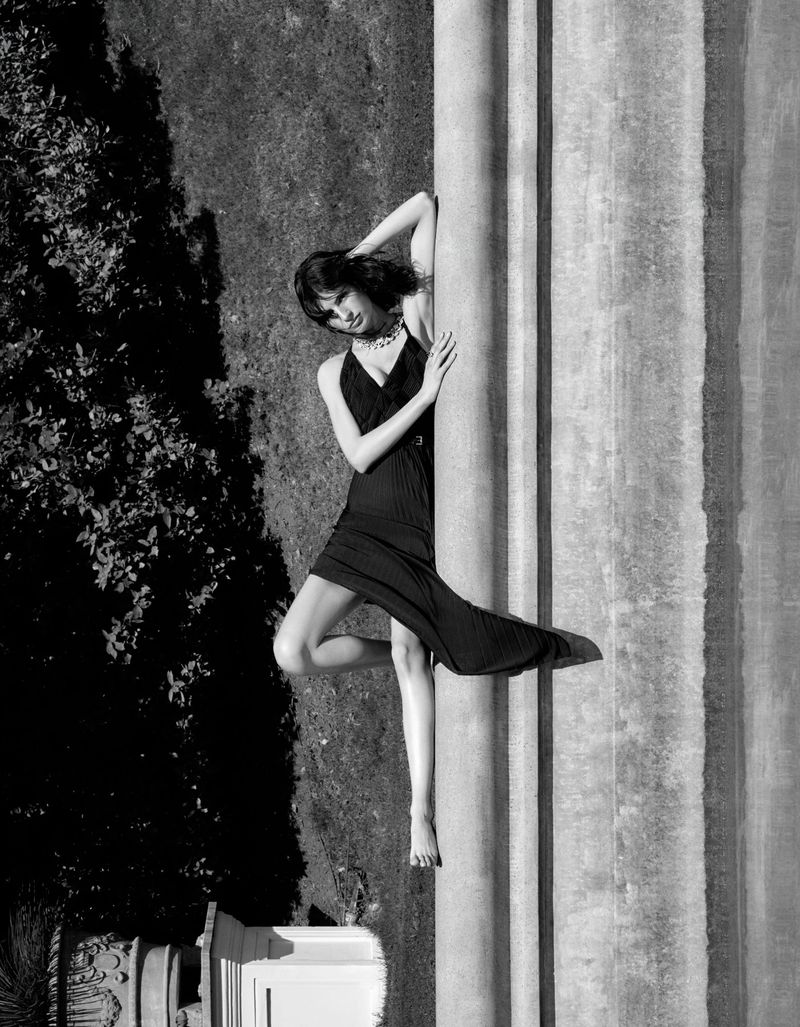 Black-and-white photo of an adult female model posing barefoot next to a pillar outdoors in a dark dress with a green grassy backdrop.