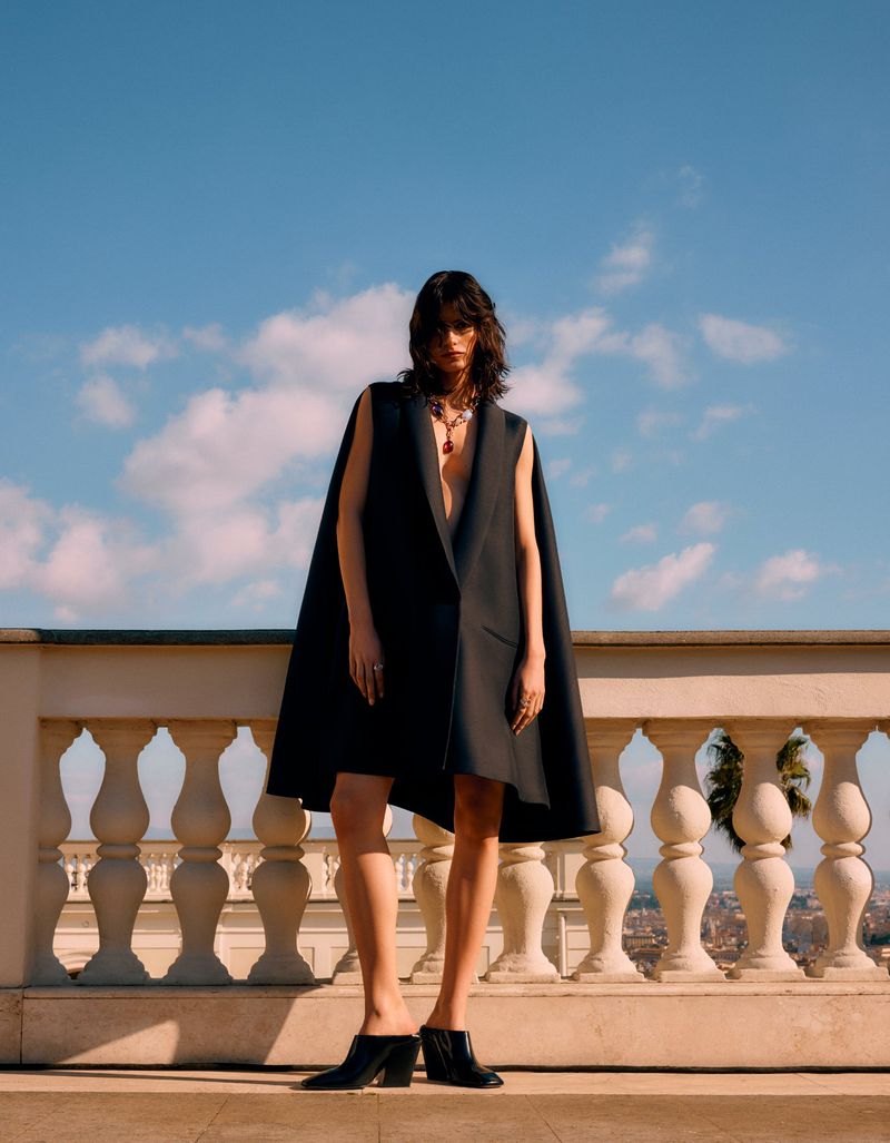 A woman in a stylish black cape dress poses outdoors against a blue sky, showcasing high fashion and elegant design on a sunny day.