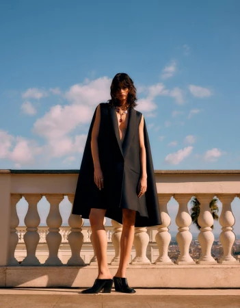 A woman in a stylish black cape dress poses outdoors against a blue sky, showcasing high fashion and elegant design on a sunny day.