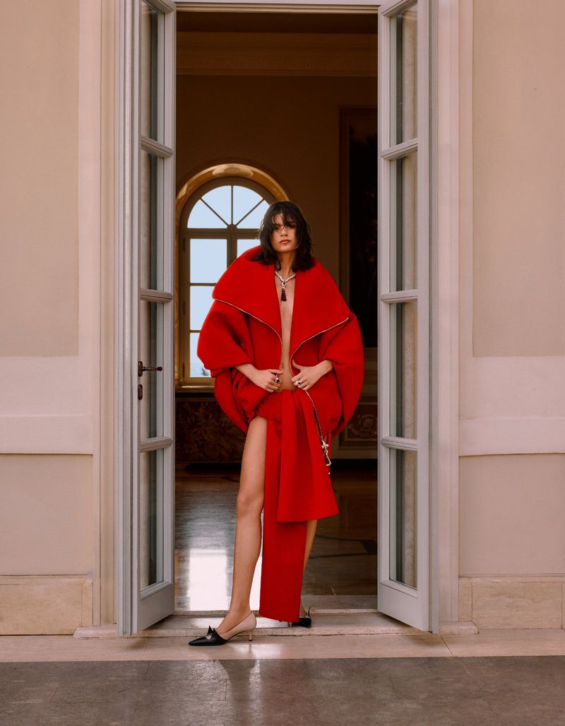 A female model posing in a red outfit stands in a doorway with natural light in an editorial fashion photograph.