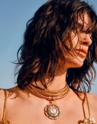 Close-up profile of a woman with wet dark hair wearing a multi-strand gold necklace against a clear sky background.