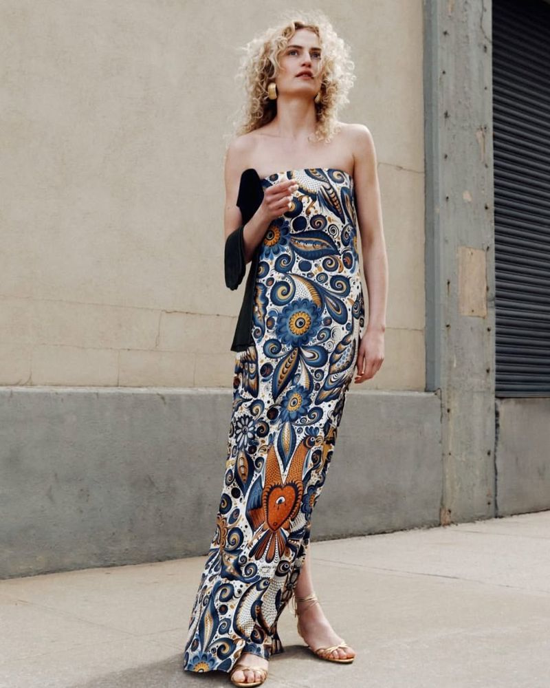 A slender woman with curly blonde hair is posing outdoors wearing a long, strapless floral dress, gold earrings, and strappy high-heeled sandals.