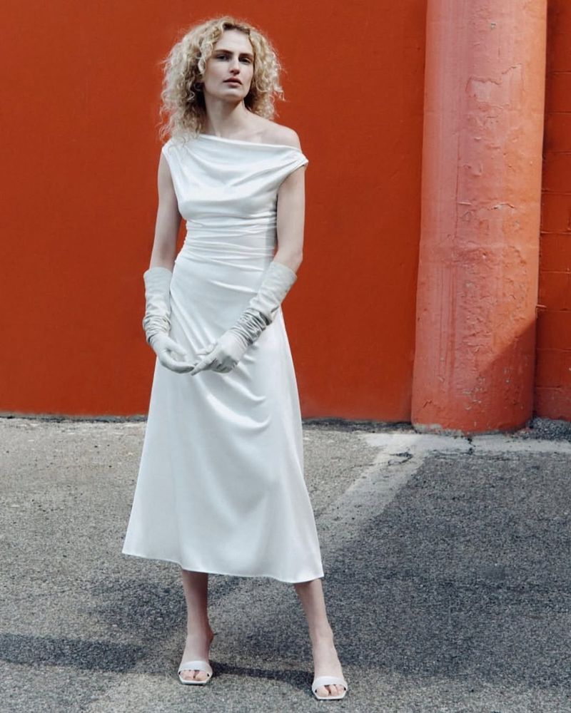 Adult female in a long white gown and light gloves poses in front of an orange wall on an overcast day in an outdoor urban setting.