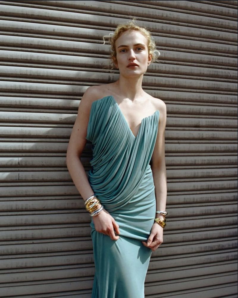 A woman with blonde curly hair wearing a blue strapless gown with gold bracelets posing in front of a gray metal roll-up door.