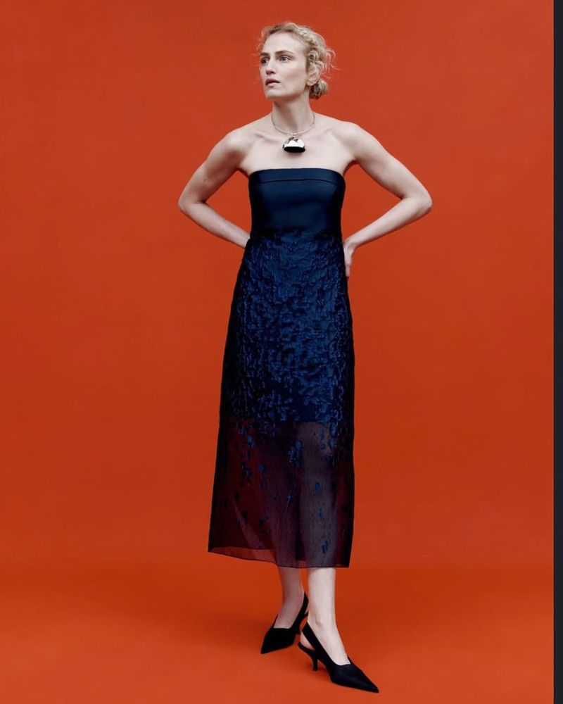 Fashionable woman in elegant navy blue dress and black heels poses against an orange backdrop, showcasing modern sophistication and style.