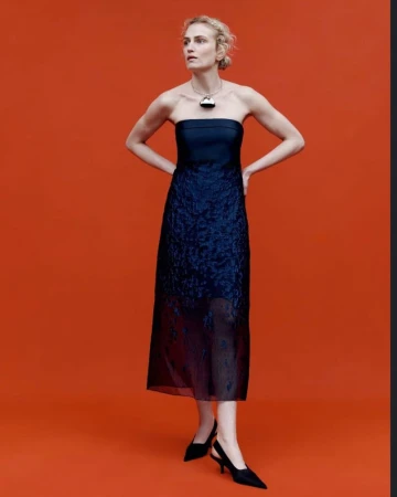 Fashionable woman in elegant navy blue dress and black heels poses against an orange backdrop, showcasing modern sophistication and style.