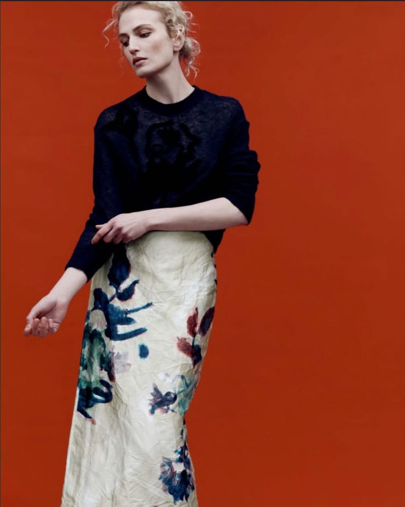 Fashion photo featuring a single adult female model wearing a black sweater and floral-print skirt against a solid orange backdrop.