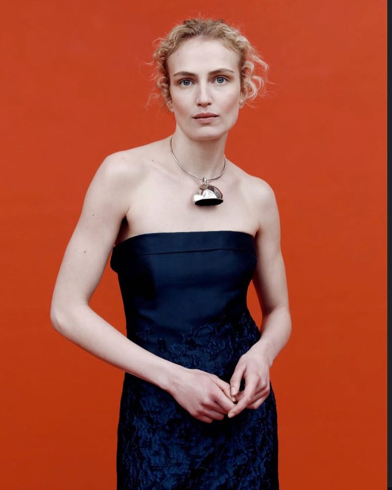 A fair-skinned fashion model with curly blonde hair wearing a strapless navy dress and necklace against an orange background.