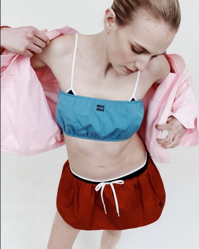 A fair-skinned adult woman poses in a light pink jacket, blue top, and red skirt in a studio shot. Miu Miu logo is visible on the top.