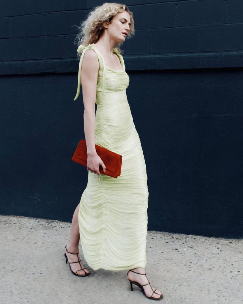 A blonde woman models a ruched yellow dress and strappy heels, carrying a clutch against a dark blue wall in an urban setting.