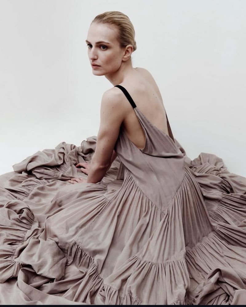 A fair-skinned woman with blonde hair in a low bun poses in a photo studio wearing a tan flowing ruffled dress with black straps.