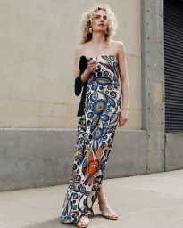Model with blonde curly hair wearing a strapless patterned blue and white dress standing outdoors on a sidewalk