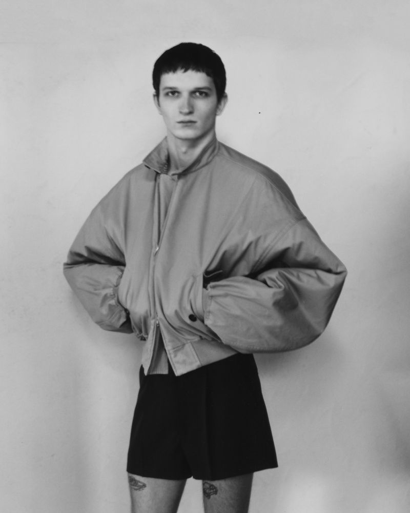 Monochrome portrait of a young adult male in a light-colored jacket and dark shorts standing against a neutral-toned wall in a studio.