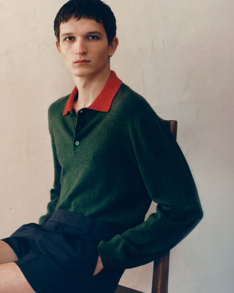 Portrait of a young adult man in a green sweater with red collar sitting indoors looking at the camera with a neutral expression.