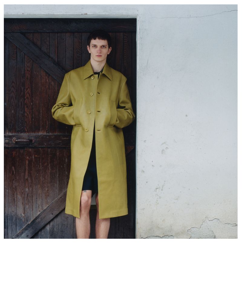 Male fashion model poses in yellow trench coat, standing against rustic door and white wall, for an outdoor fashion editorial.