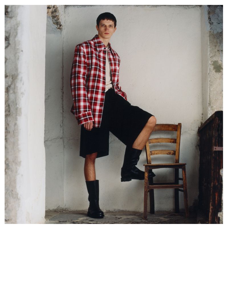 A young man in a plaid shirt and black shorts poses with his leg on a wooden chair indoors, looking at the camera with a neutral expression.