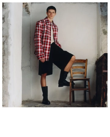 A young man in a plaid shirt and black shorts poses with his leg on a wooden chair indoors, looking at the camera with a neutral expression.