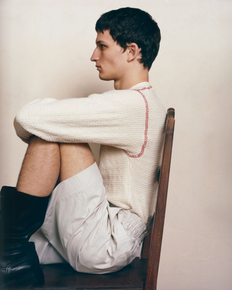 Profile shot of a young man in a cream sweater and light shorts sitting on a wooden chair against a neutral background in studio lighting.