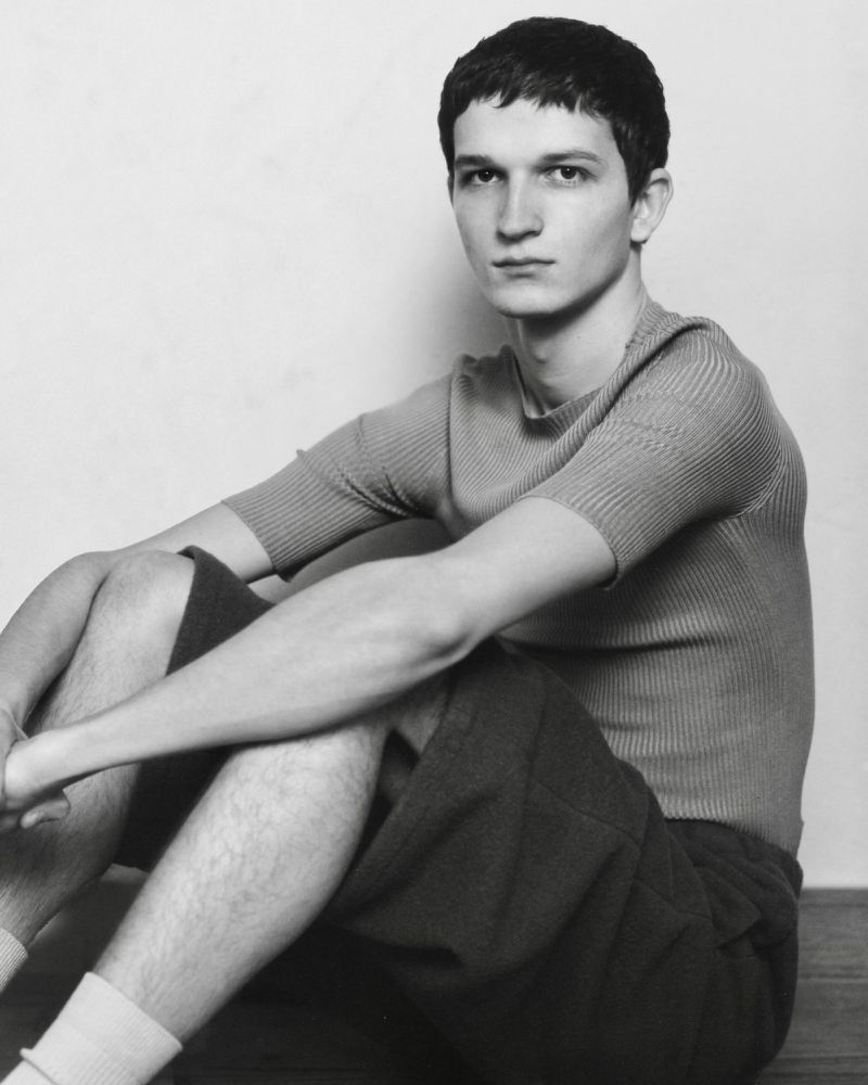 Black and white portrait of a young adult man sitting indoors. He wears a short-sleeved shirt and shorts. Neutral expression, minimal background.