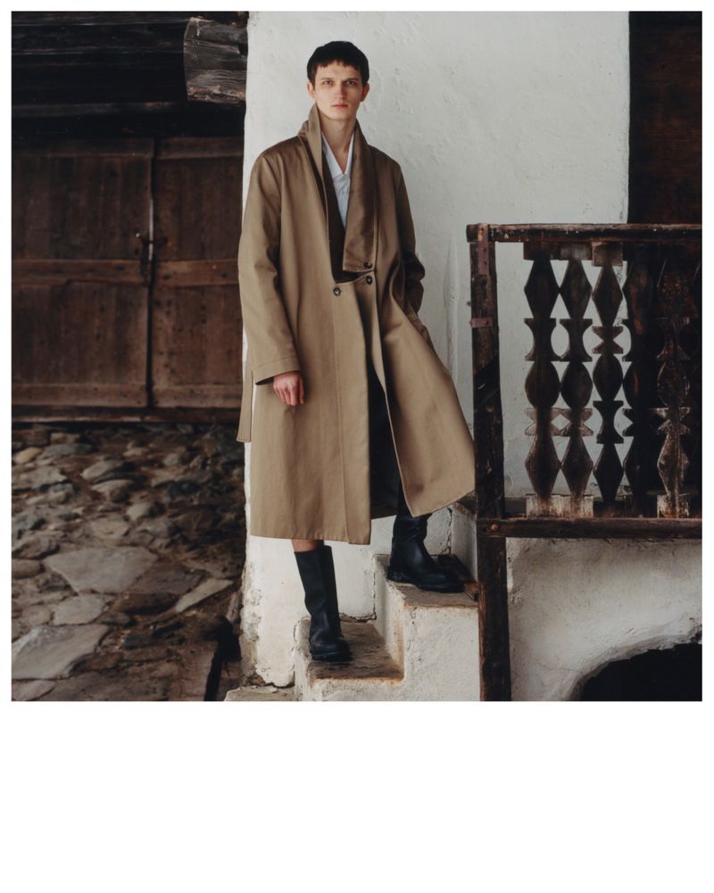 A young adult poses on weathered steps wearing a long tan coat and boots against a rustic white stone wall with a weathered wooden door.