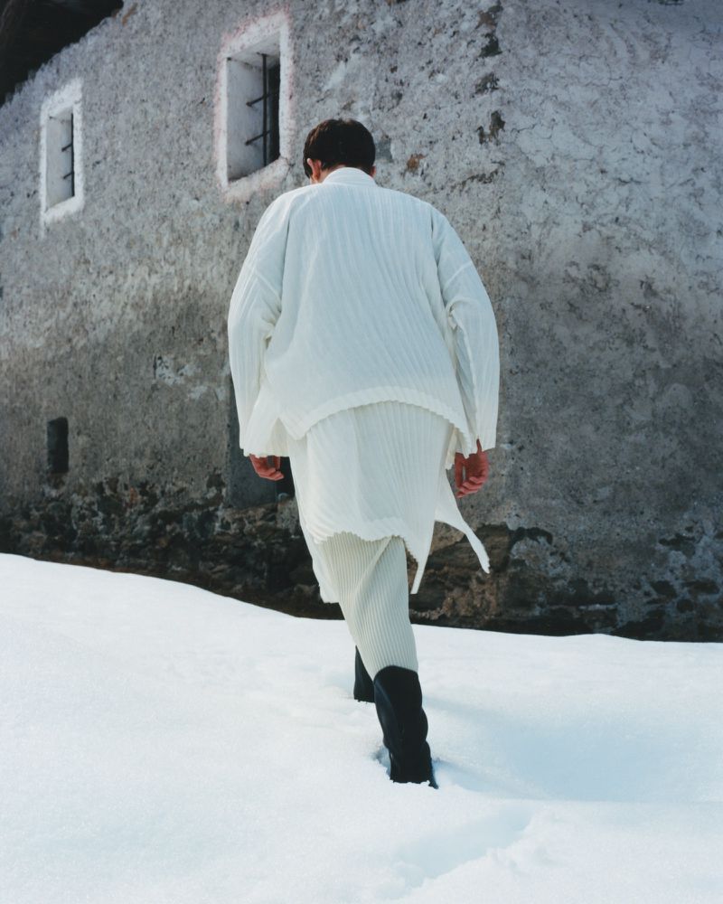 An adult is walking away in the snow wearing white clothing and black boots, with an old grey building in the background.