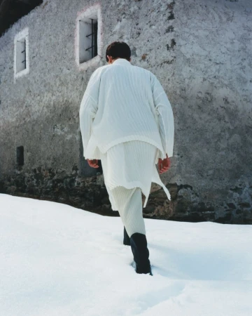 An adult is walking away in the snow wearing white clothing and black boots, with an old grey building in the background.