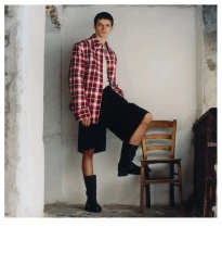A male model wearing a red plaid jacket and black shorts standing with one foot on a wooden chair.