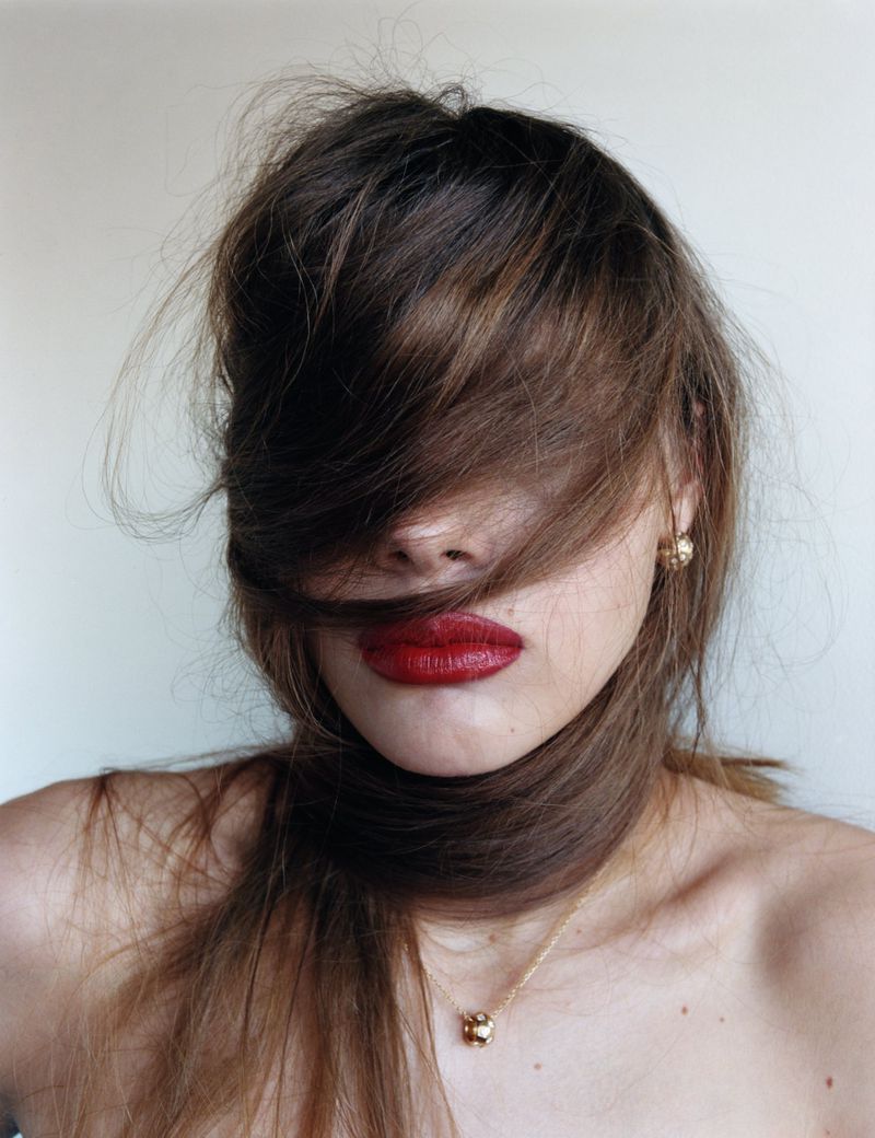 Close-up portrait of a woman with brown hair partially covering her face, red lipstick, gold earrings, and a gold necklace.