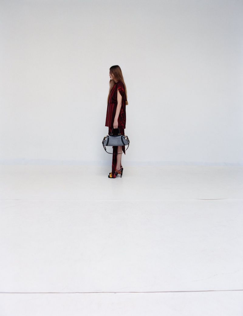 Adult female poses in a photo studio. She wears a maroon dress and patterned high heels, and carries a light blue purse.