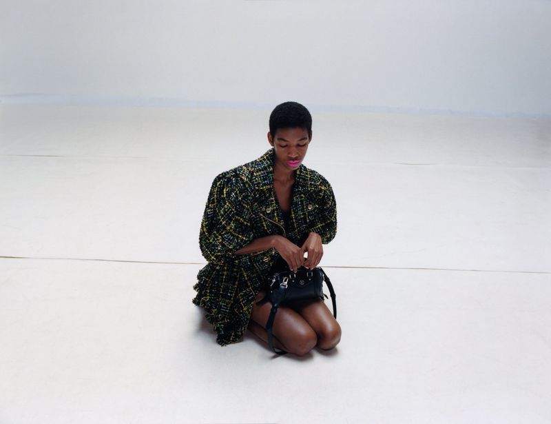 A young Black woman in a patterned jacket kneels with a black purse against a bright, neutral background in a fashion studio setting.