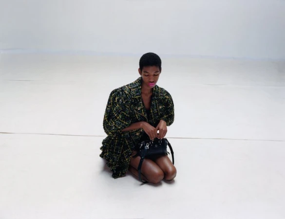 A young Black woman in a patterned jacket kneels with a black purse against a bright, neutral background in a fashion studio setting.