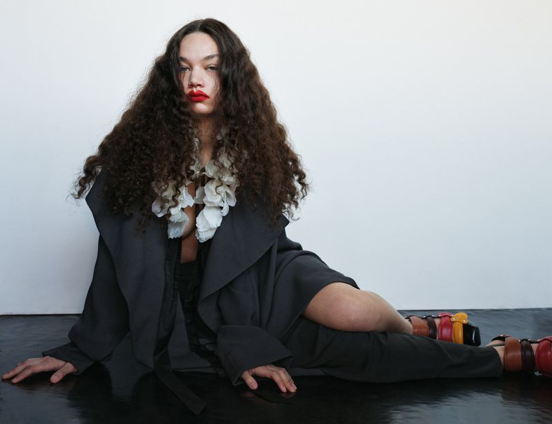 Woman with curly brown hair and red lipstick poses while wearing a black coat and colorful platform sandals on a glossy black surface.