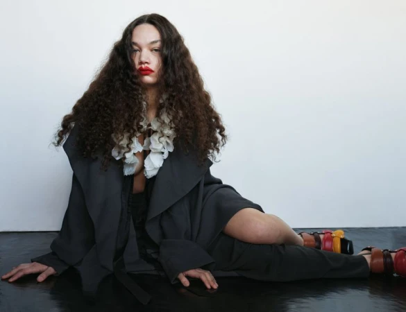 Woman with curly brown hair and red lipstick poses while wearing a black coat and colorful platform sandals on a glossy black surface.