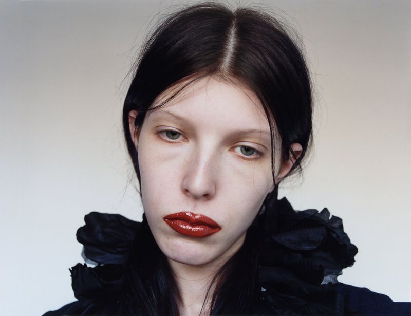 Close-up portrait of a young adult female with black hair, wearing a black ruffled top and red lipstick, against a light gray background.