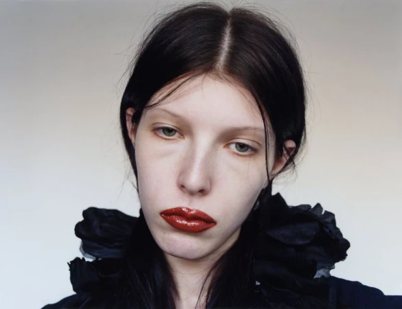 Close-up portrait of a young adult female with black hair, wearing a black ruffled top and red lipstick, against a light gray background.