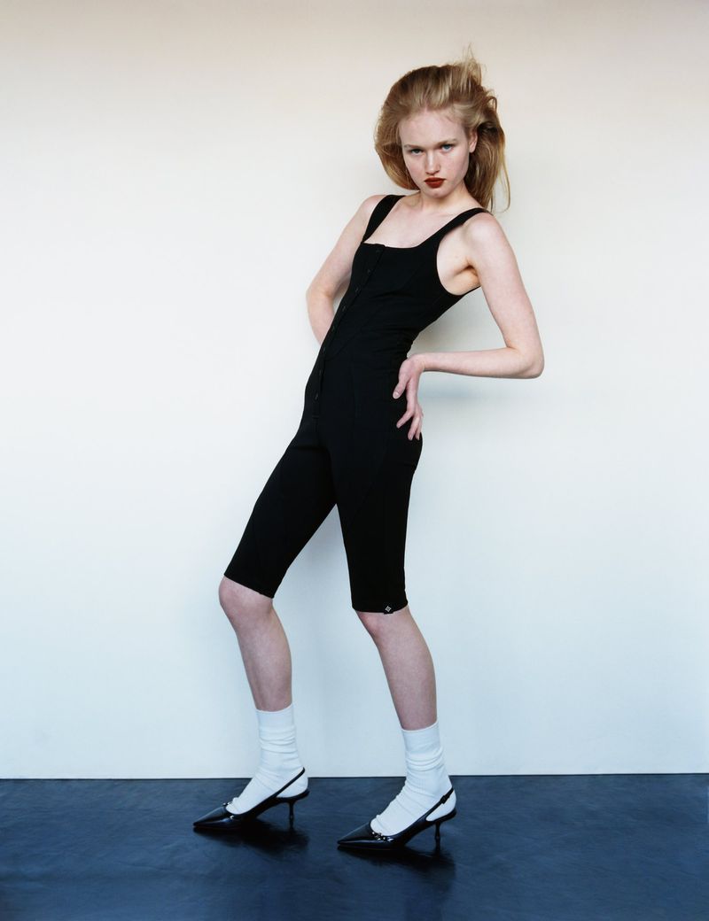A slender woman in a black playsuit poses against a neutral backdrop, her dark red lipstick accentuating her features, fashion.