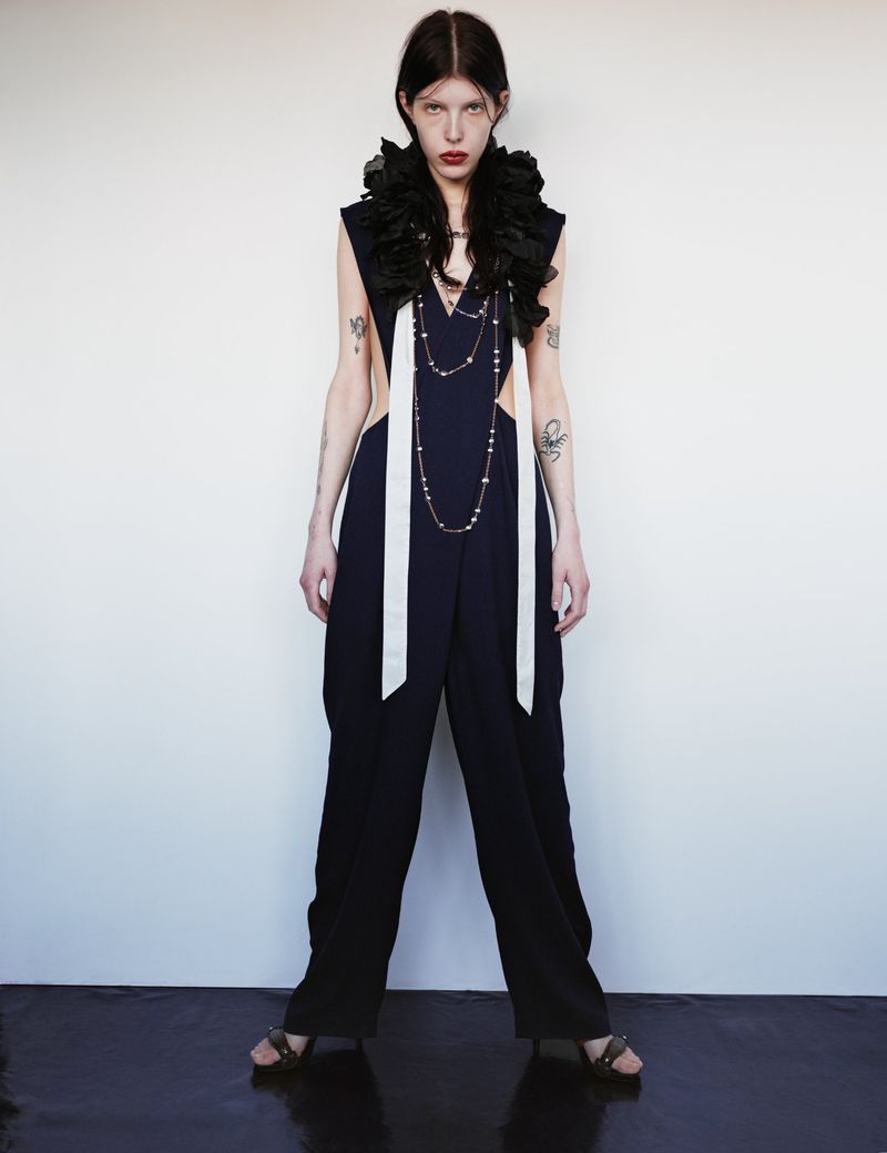 Fashion-forward editorial shot of a slender woman in a dark jumpsuit with white details and a dramatic black ruffle collar against a white wall.