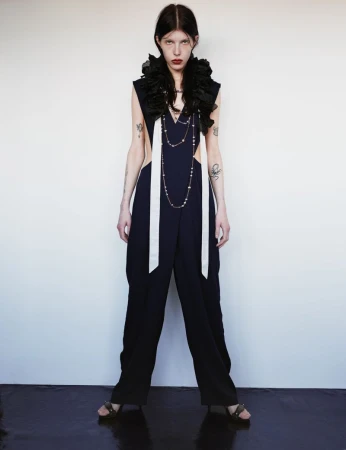 Fashion-forward editorial shot of a slender woman in a dark jumpsuit with white details and a dramatic black ruffle collar against a white wall.