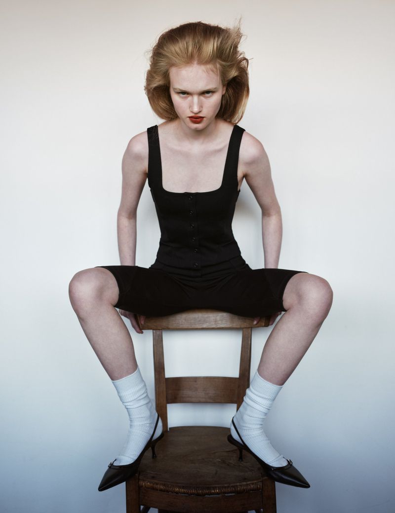 Portrait of a young woman with red hair sitting on a wooden chair in a black outfit, white socks, and black high heels against a white background.