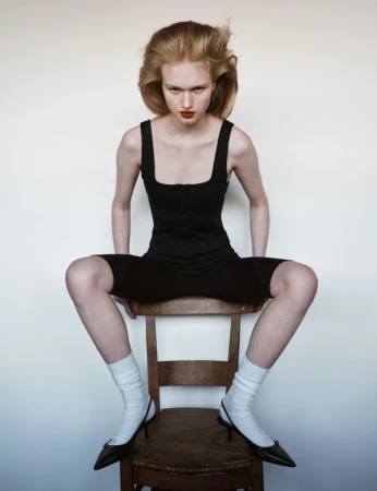 Portrait of a young woman with red hair sitting on a wooden chair in a black outfit, white socks, and black high heels against a white background.